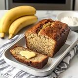 A loaf of banana bread with a slice topped with butter, alongside ripe bananas and a bowl of cream, on a kitchen countertop.