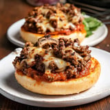 Two toasted English muffins topped with ground beef, mushrooms, marinara sauce, and melted cheese, served on white plates.