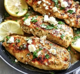 A close-up image of seasoned grilled chicken breasts garnished with feta cheese and fresh herbs, served with lemon slices.