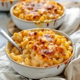 A close-up of a bowl of cheesy, baked macaroni and cheese, with a crispy golden top and a spoon resting beside it.