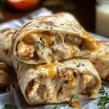 A close-up of a cheesy, chicken-filled burrito, partially opened to reveal melted cheese and chopped herbs, resting on parchment paper, accompanied by some nuts.