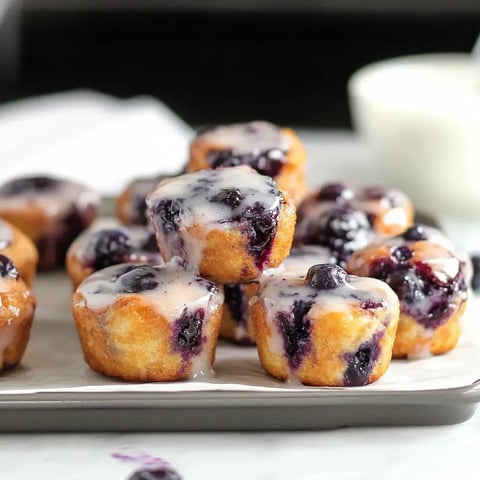 A close-up of golden-brown mini muffins topped with a glossy glaze and scattered blackberries.