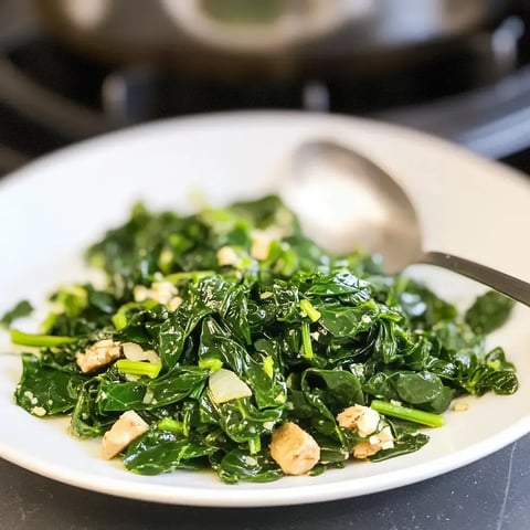 A white plate filled with cooked, seasoned leafy greens and chopped pieces of chicken, with a spoon resting beside it.